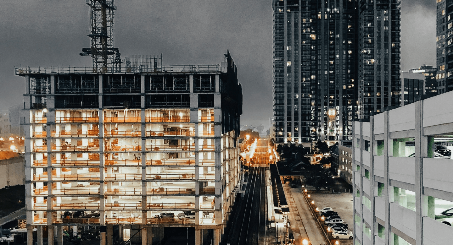 A high-rise under construction at dusk, its unfinished concrete floors lit from within, with a railway line running past the site.